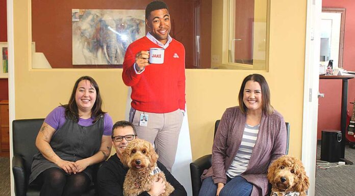 State Farm agent offers insurance plus puppy love! Surrounding a cardboard cutout of “Jake,” State Farm Insurance’s chief advertising spokesperson, are Mark Creevey’s office “staff,” (from left) Gillian Moreno, Creevey holding his golden-doodle Willow, Sarah Kimsey-Sauro and visiting golden-doodle Olive.
