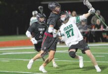 Tigard lacrosse teams see growth despite records A Tigers lacrosse player in a white jersey (number 27) maneuvers past two Tualatin defenders in black jerseys during a night game on a turf field.