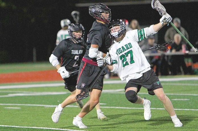 A Tigers lacrosse player in a white jersey (number 27) maneuvers past two Tualatin defenders in black jerseys during a night game on a turf field.