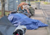 City cracks down on ‘camp materials’ in city rights-of-way A sidewalk in Tigard is partially blocked by a collection of personal items and camping gear, including multiple blue tarps and a portable toilet in the background. A person and a dog are visible amidst the belongings.