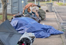 City cracks down on ‘camp materials’ in city rights-of-way A sidewalk in Tigard is partially blocked by a collection of personal items and camping gear, including multiple blue tarps and a portable toilet in the background. A person and a dog are visible amidst the belongings.