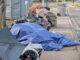 City cracks down on ‘camp materials’ in city rights-of-way A sidewalk in Tigard is partially blocked by a collection of personal items and camping gear, including multiple blue tarps and a portable toilet in the background. A person and a dog are visible amidst the belongings.