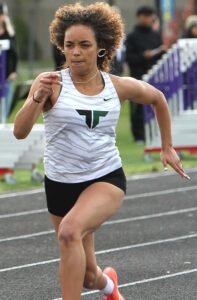Tigard High School track athlete Sydney Swayne sprinting at full speed in a white tiger-striped jersey during a 100-meter dash.
