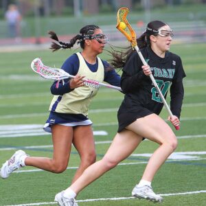 A Tigard High School girls' lacrosse player in a black jersey (number 21) cradles a yellow ball in her stick while running down the field, closely pursued by a defender in a gold jersey.