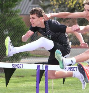 Tigard High School athlete Brady Blanchard in mid-air clearing a white hurdle with "SUNSET" printed on it during a track and field race.