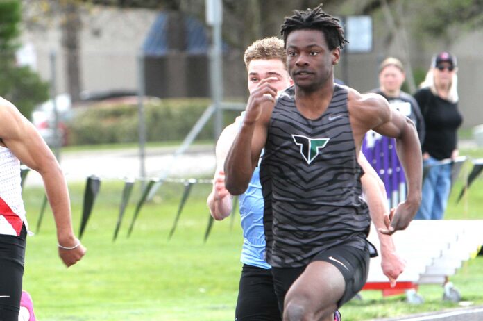Tigard track athlete Marcus Burton, wearing a grey tiger-striped jersey, sprinting at full speed during a 100-meter dash on an outdoor track.